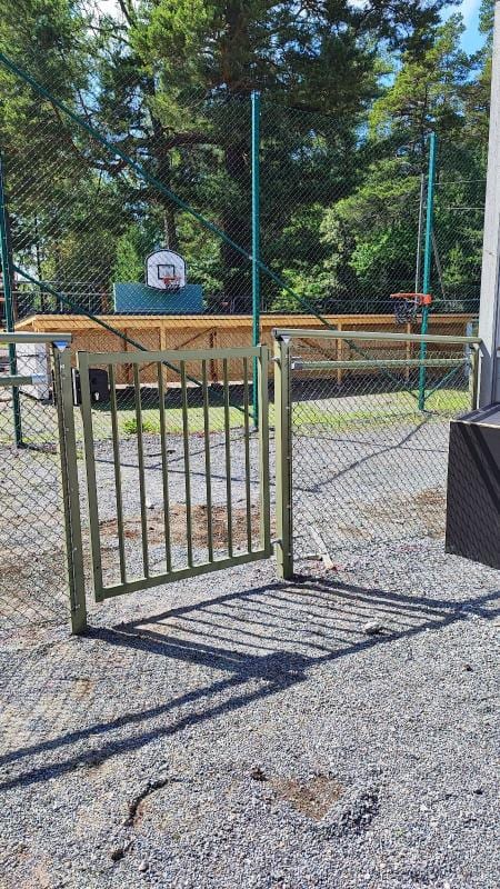 Gånggrind i stål med flätverksstängsel runt en bollplan.